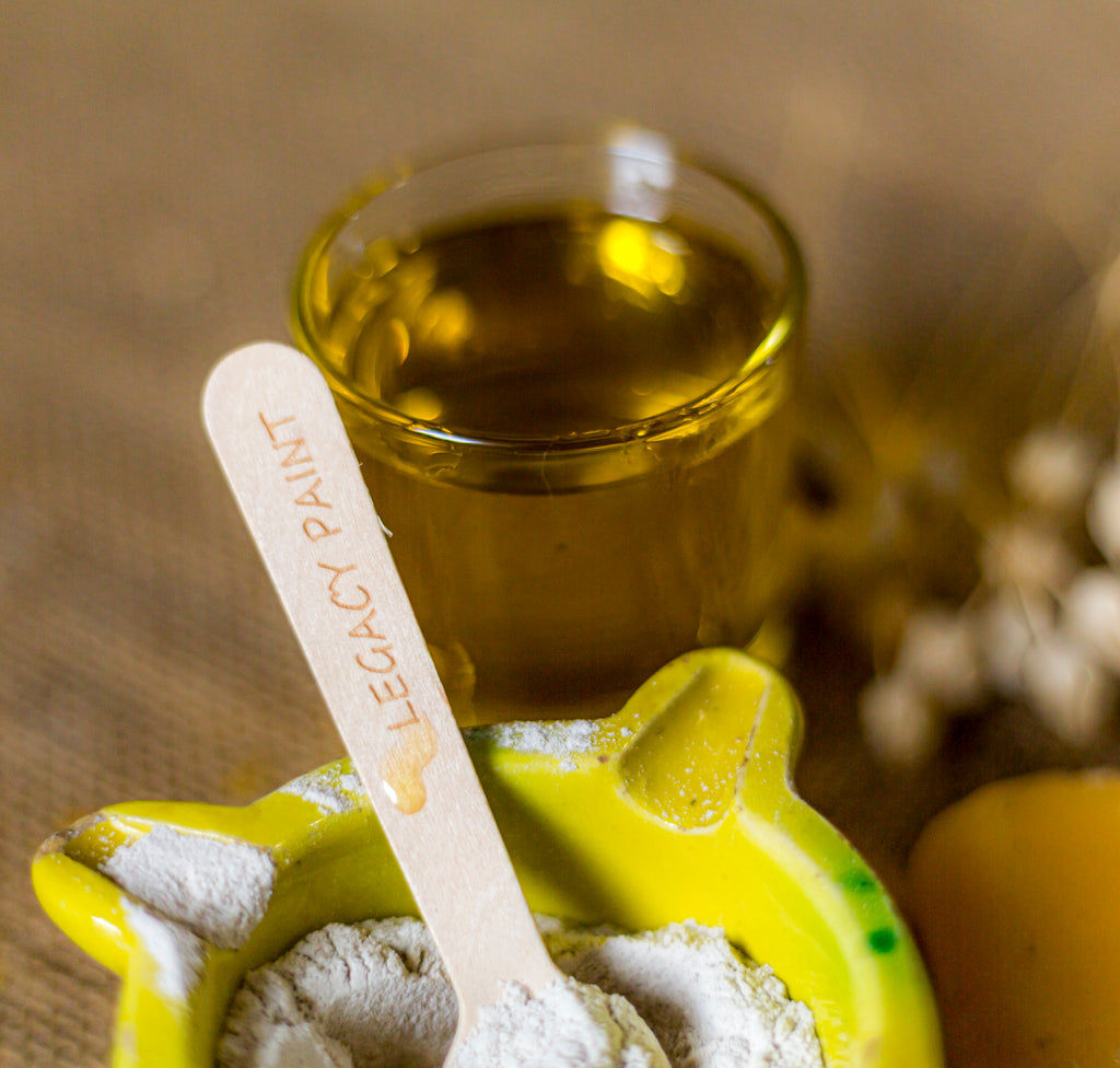 Legacy Paint Artisan Alchemy: A traditional Spanish mortar with raw Spanish White and a droplet of pure Spanish olive oil on a branded wooden spoon. This image captures the raw mineral composition of our encaustic-based waxes, highlighting the organic Spanish matter and ancestral tools used by Legacy Paint for bespoke furniture finishes."